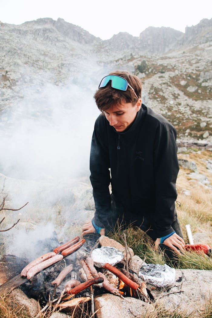A young man prepares sausages on a campfire amid the mountainous landscape of Barèges, France.