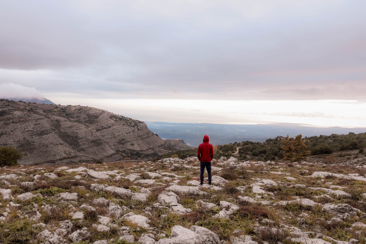 Person in red hoodie standing alone on a rocky terrain with mountains in the distance.
